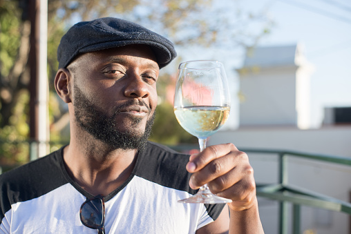 Close-up of happy African man holding glass of wine. Young handsome man in black cap tasting drinking wine looking at camera on city and blue sky background. Celebration, dating, happy moments concept
