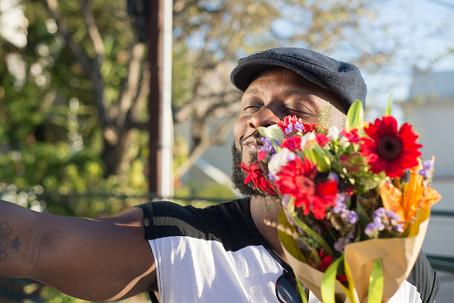 Portrait of happy African American man with bunch of flowers. Young bearded man in black cap holding beautiful flowers posing enjoying warm sunny weather with closed eyes. Happiness, dating concept