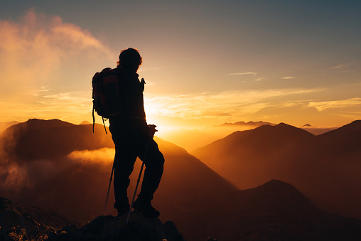 backlit silhouette of hiker with backpack and poles on top of a mountain watching the sunset. sport and healthy life concept.