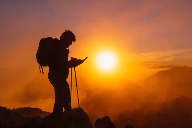 backlit silhouette of hiker with backpack using gps apps on his mobile phone