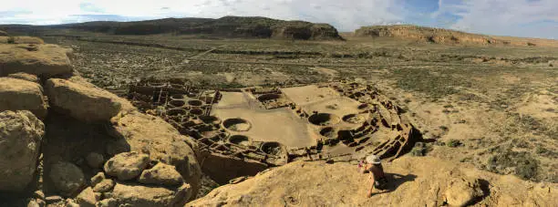 In the morning, a woman after a hike sits down to enjoy an overlook view of Pueblo Bonito, a Chocoan great house of Ancestral Pueblo dwellings in New Mexico containing over 600 rooms and occupied from 828 A.D. and 1126 A.D. in Chaco Culture National Historical Park.