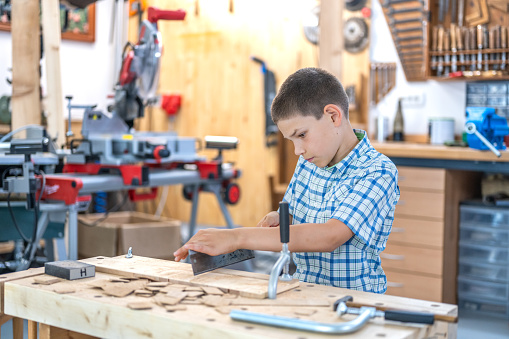 Boy cutting wooden plank, holding handsaw in carpentry workshop, medium shot. Child working with woodwork tool at art class for kids