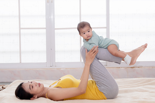 Beautiful Asian mother and daughter doing yoga exercise home. Young mom lift infant up playing airplane on bed. Happy mother lifting baby in air. Woman and toddler play together with love and caring.