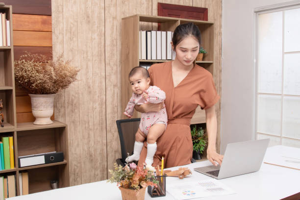 Beautiful Asian mother holding todlder and trying to work from home on laptop, mom works at computer with child. Newborn in pink cotton soft textile cherry pattern. Childcare and working mom concept. hermosa madre asiática sosteniendo a todlder y tratando de trabajar desde casa en una computadora portátil, mamá trabaja en la computadora con el niño. recién nacido en algodón rosa suave textil patrón cereza. concepto de cuidado de niños y madres