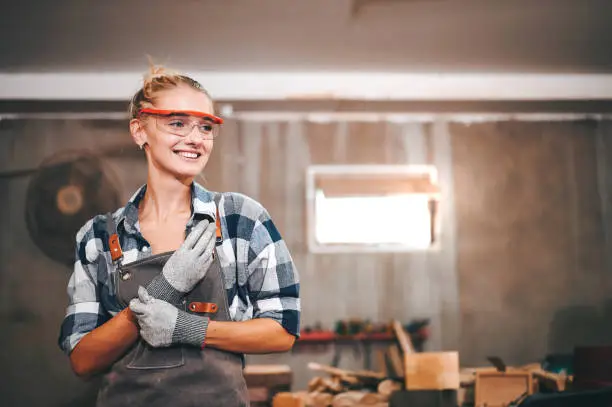 Carpenter woman wearing gloves and eye protection standing smile in wood workshop.