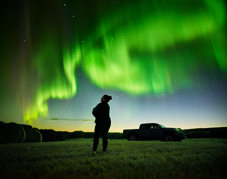 Silhouette of a woman standing in farm field and looking at northern lights in the night sky
