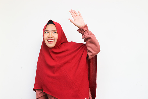 Beautiful asian muslim in hijab looking up and wave hand to greeting, isolated on white background