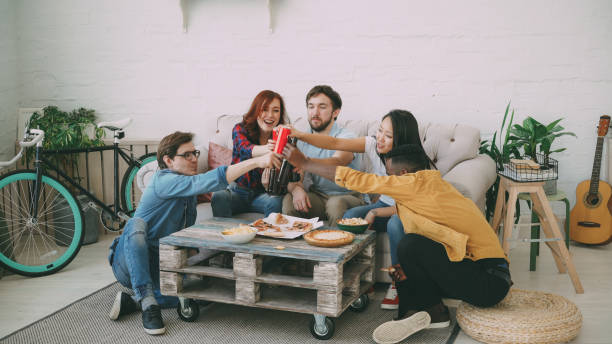 Happy friends have party talking and eating pizza. They clink bottles sitting on sofa at home Happy friends have party talking and eating pizza. They clink bottles sitting on sofa at home indoors roommate stock pictures, royalty-free photos & images