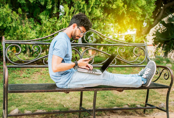 Freelancer man sitting in a park using laptop and cellphone. Man in a park working online with laptop. Relaxed man working with laptop outdoor, Young man in a park with laptop and cell phone hombre independiente sentado en un parque usando computadora portátil y teléfono celular. hombre en un parque trabajando en línea con computadora portátil. hombre relajado trabajando con computadora portátil al aire libre, joven en un parque con compu