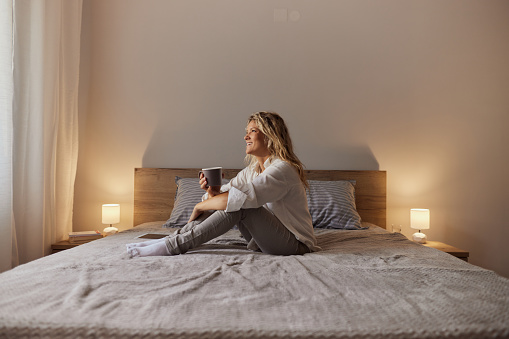 Young happy woman day dreaming while drinking coffee and relaxing on a bed. Copy space.