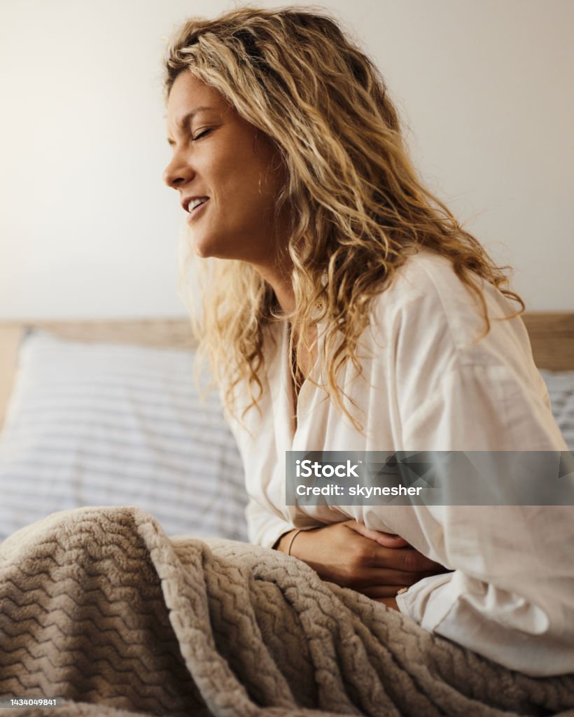 Ugh, I have a stomachache! Young woman holding her stomach in pain while relaxing on a bed. Home Interior Stock Photo