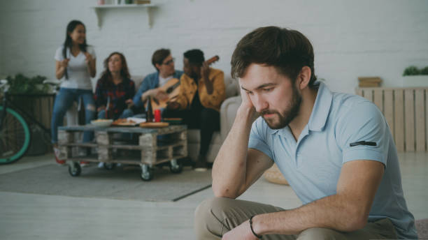 young student guy feels upset and isolated while his friends celebrating party at home indoors - solidão imagens e fotografias de stock