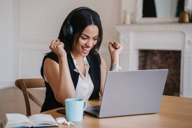 Beautiful African american young woman in headphones squishing fist like winner smiling, making distant call by laptop , working from home. Brazilian happy girl call center employe celebrates deal. hermosa joven afroamericana con auriculares apretando el puño como ganadora sonriendo, haciendo llamadas lejanas por computadora portátil, trabajando desde casa. el empleado del centro de llamadas de la chica feliz brasileña celebra el trato.