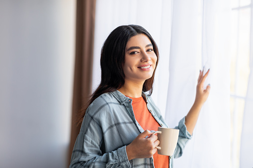 Happy young Arab lady drinking fresh coffee near window at home, enjoying relaxing morning. Portrait of middle Eastern woman holding cup of hot aromatic beverage indoors