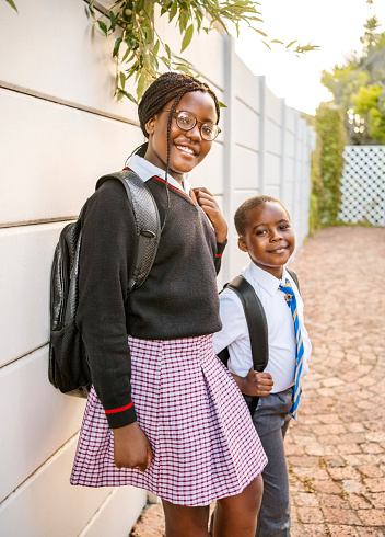 Portrait of a smiling young brother and sister in school uniform standing outside of their home