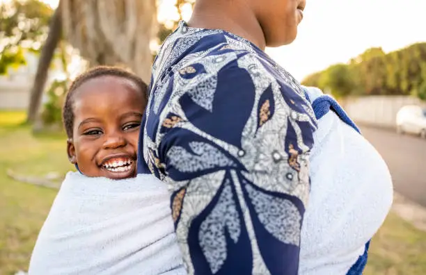 Close-up of cute boy sitting back wrap sling of his mother walking outside