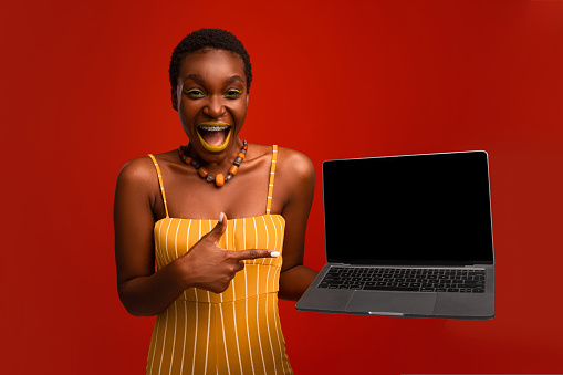 Emotional stylish african american young woman in bright outfit showing brand new laptop with empty screen, posing on red studio background, showing nice website or software, mockup