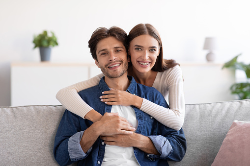 Happy young wife hugs husband from behind on sofa and look at camera in room interior, empty space. Love, happiness, relationship. Family enjoy time together, weekend at home during covid-19 outbreak