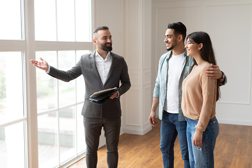 Portrait Of Male real estate agent Or Architect Wearing Suit Showing View From New Empty House To Smiling Millennial Buyers, Pointing At Window. Husband Hugging Wife, Visiting Residential Building Choosing Home