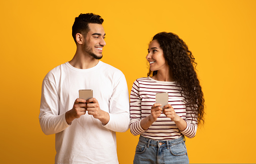 Mobile Communication. Cheerful Arab Man And Woman Holding Smartphones And Looking At Each Other, Happy Middle Eastern Couple Using Cellphones While Standing Together On Yellow Background, Copy Space