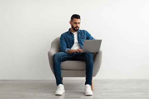 Full length of millennial Arab man using laptop for online work, studies or communication, sitting in armchair against white studio wall. Young middle Eastern guy browsing web on portable pc