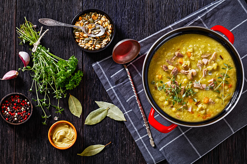 Finnish split pea soup Hernekeitto with ham on red pot with hot mustard and pumpkin seeds on dark wood table, horizontal view from above, flat lay