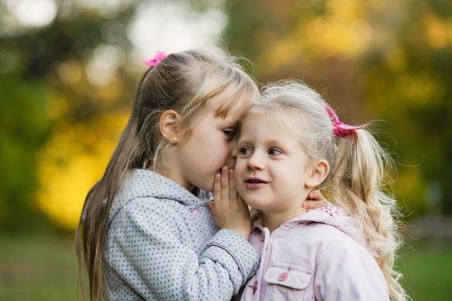 A little girl tells the secret in her friend's ear. Two cute baby girls in the autumn park.