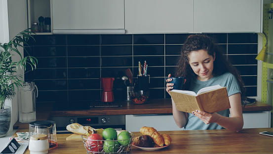Beautiful curly woman sitting at table holding cup of coffee, drinking and reading book in nice modern kitchen at home, she is positive and smiling