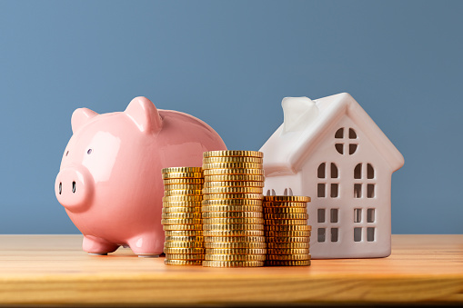 Savings: Front view of a wooden table with a piggy bank, a stack of coins and a house shape against a dark blue wall.