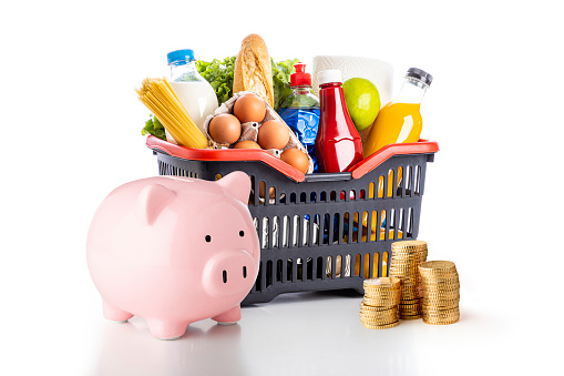 Front view of a piggy bank beside a sopping basket full of groceries and a coin stack isolated on white background