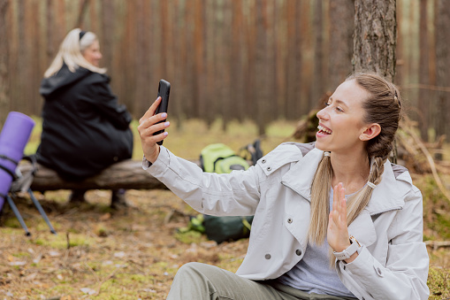 Satisfied woman with blonde hair tied in braids sits on the ground in the forest talking on the phone video chat waving to the camera, resting while trekking, camping, hiking.