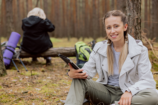 Young daughter and elderly mom spending time together in woods hikking smiling young woman in the foreground holding tablet sitting on the ground looking at camera and elderly woman in the background.
