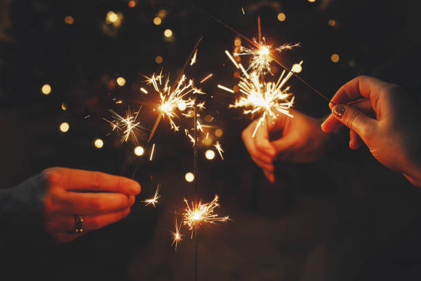 ¡feliz año nuevo! amigos celebrando con bengalas encendidas en las manos contra las luces del árbol de navidad en el cuarto oscuro. manos sosteniendo fuegos artificiales sobre el fondo de un elegante árbol iluminado decorado. malhumorado - new years fotografías e imágenes de stock