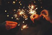 Happy New Year! Friends celebrating with burning sparklers in hands against christmas tree lights in dark room. Hands holding fireworks on background of stylish decorated illuminated tree. Moody