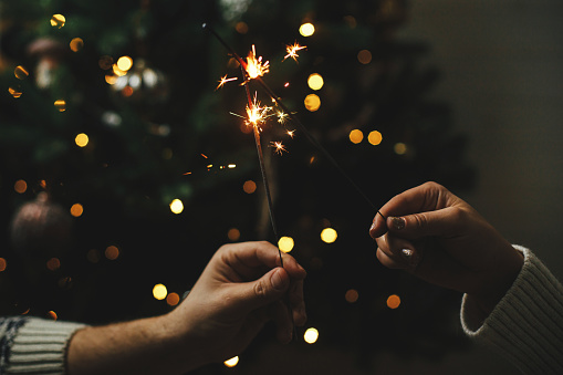 Happy New Year! Couple celebrating with burning sparklers in hands on background of stylish decorated tree with illumination. Hands holding firework against christmas tree lights in dark room