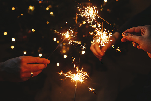 Happy New Year! Hands holding fireworks against christmas lights in dark room. Atmospheric holiday. Friends celebrating with burning sparklers in hands on background of stylish illuminated tree