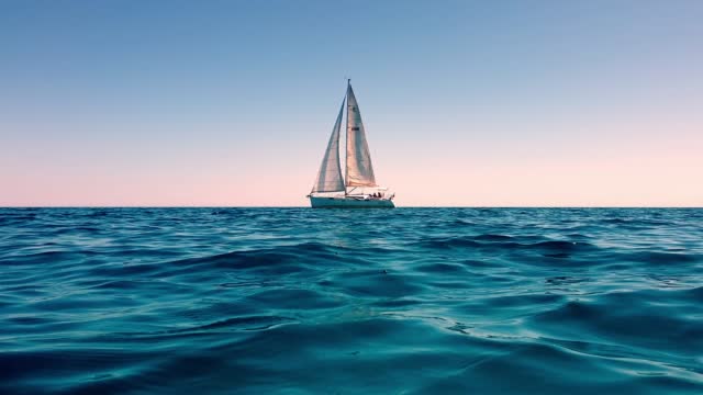 Slow-motion and low-angle sea-level view of small yacht boat sailing in calm open sea at sunset