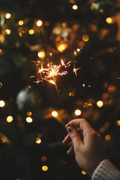 frohes neues jahr!! brennende wunderkerze in weiblicher hand auf dem hintergrund von weihnachtsbaumlichtern in dunklem raum. stimmungsvolle feier. hand hält feuerwerk gegen stilvoll geschmückten baum mit beleuchtung - sparkler sparks new years eve human hand stock-fotos und bilder