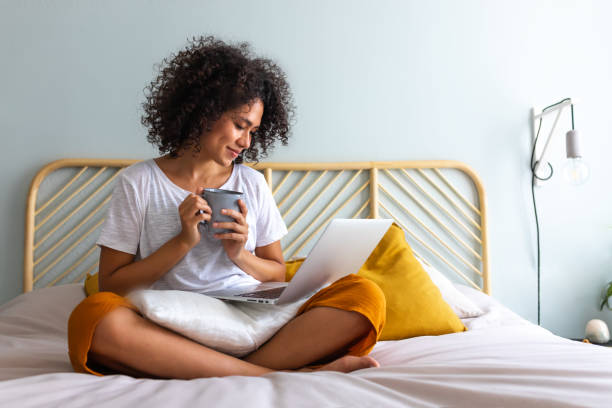 istock Cozy at home. African American woman sitting on bed drinking coffee while watching show on laptop. Relaxing in bedroom. 1434091061