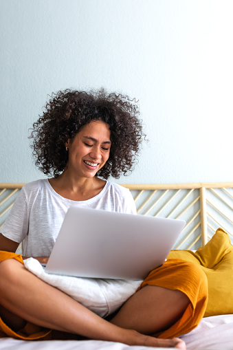 Smiling young multiracial latina woman with curly hair using laptop sitting on bed at home. Vertical image. Copy space. Technology concept.