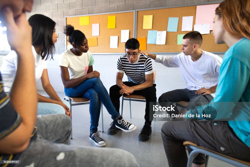 Sad hispanic male high school student sharing bad news with classmates. Multiracial teenagers sitting in circle in Sad hispanic male high school student sharing bad news with classmates. Multiracial teenagers sitting in circle in class comfort depressed teen boy. Mental health and education concepts. Mental Health Stock Photo
