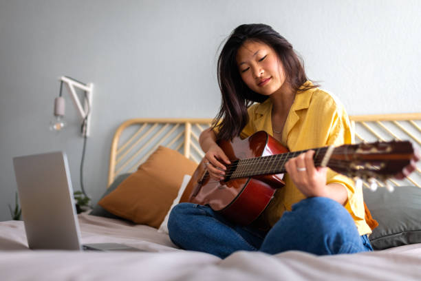 ragazza asiatica adolescente che impara a suonare la chitarra acustica seguendo lezioni di video tutorial online usando il laptop. e-learning - lyricist foto e immagini stock
