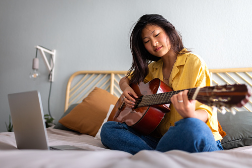 Teenage asian girl learning to play acoustic guitar following online video tutorial classes using laptop. E-learning and hobbies.