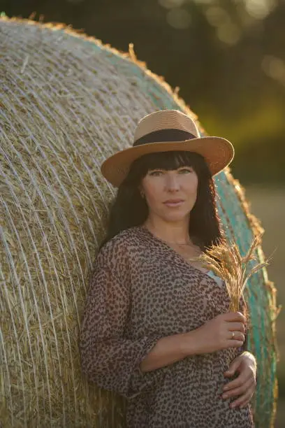 A pregnant beauty with black long hair leaned on a haystack, she was dressed in a fashionable dress, a straw hat on her head and a spikelet of grain in her hands. The joy of motherhood. Mother-to-be. Healthy childbirth.