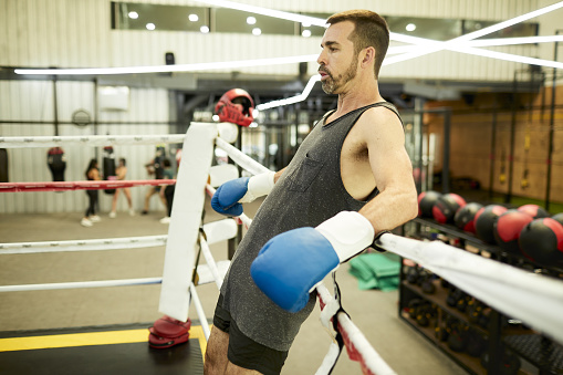 Exhausted male leaning against the boxing ropes to rest up after a sparring session. Unrecognizable women in blurred background. Three quarter length close up with copy space.