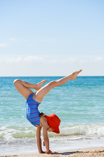 Teenage girl stay on hands with legs up at summer sea beach in swimsuit and sun hat, active happy childhood, memorable family vacation