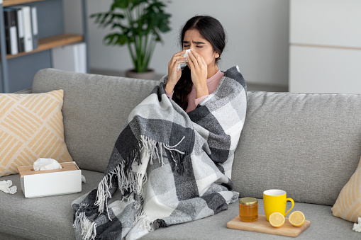 Unhappy young indian lady sneezes in napkin, blows nose, suffers from fever, flu on couch in room interior with tea, honey and lemon. Home treatment for cough, runny, cold and covid-19, self-isolation
