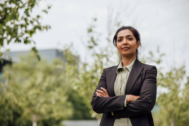 A waist-up shot of a confident businesswoman looking into the camera with her arms crossed with a serious expression on her face. She is dressed smartly in a suit jacket and standing in a business park. She is based in the North East of England