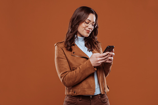 Smiling young brunette woman girl in brown jacket using mobile phone posing isolated over orange wall background studio portrait. Smartphone, typing sms message, technology, people lifestyle concept. Mock up copy space.