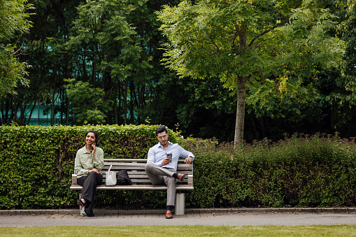 A full shot of a business male and female sitting down on a bench. They are dressed smartly in a business park. They are both on their phones whilst on a break from work. They are based in the North East of England.